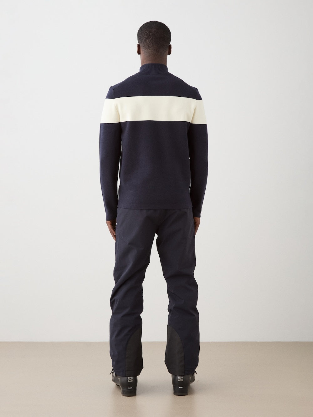 We Norwegians | FW25 | Mens Holmenkollen ZipUp_Navy blue | Model is wearing a merino/ cotton blend - knitted sweater with a 1/4th zipper opening and badge at the front - paired with black ski pants | Shot against an off-white background | Back view