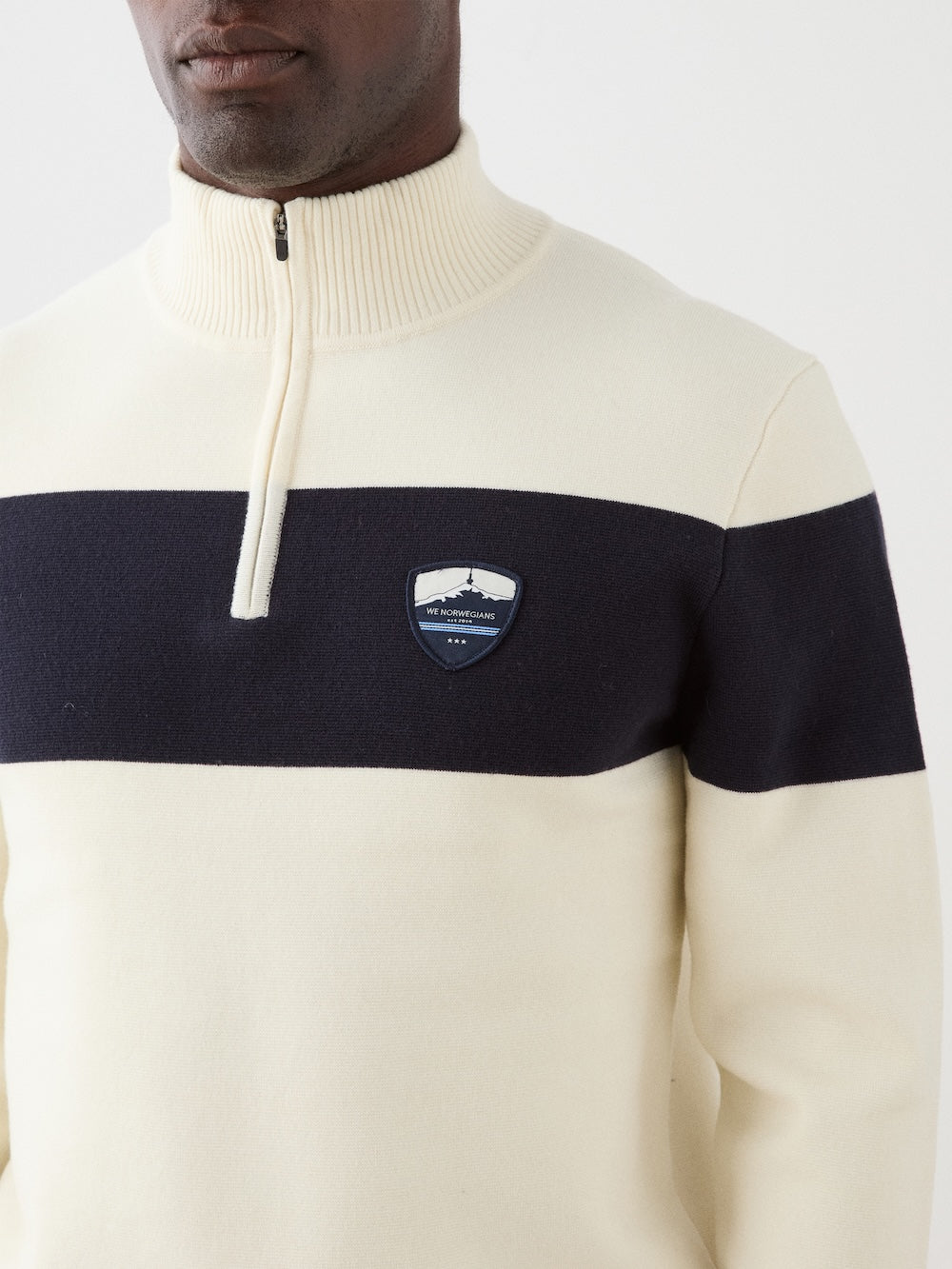 We Norwegians | FW25 | Mens Holmenkollen ZipUp_Off white | Model is wearing a merino/ cotton blend - knitted sweater with a 1/4th zipper opening and badge at the front | Shot against an off-white background | Close-up view