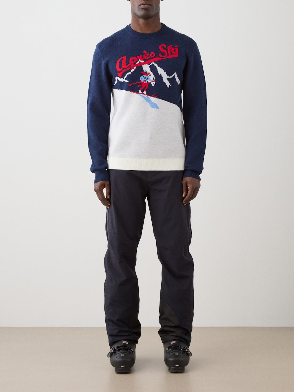 We Norwegians | FW25 | Mens Afterski Crewneck_Navy blue | Model wearing a knitted sweater with an après ski design at the front with black ski pants | Shot against an off-white background | Front view