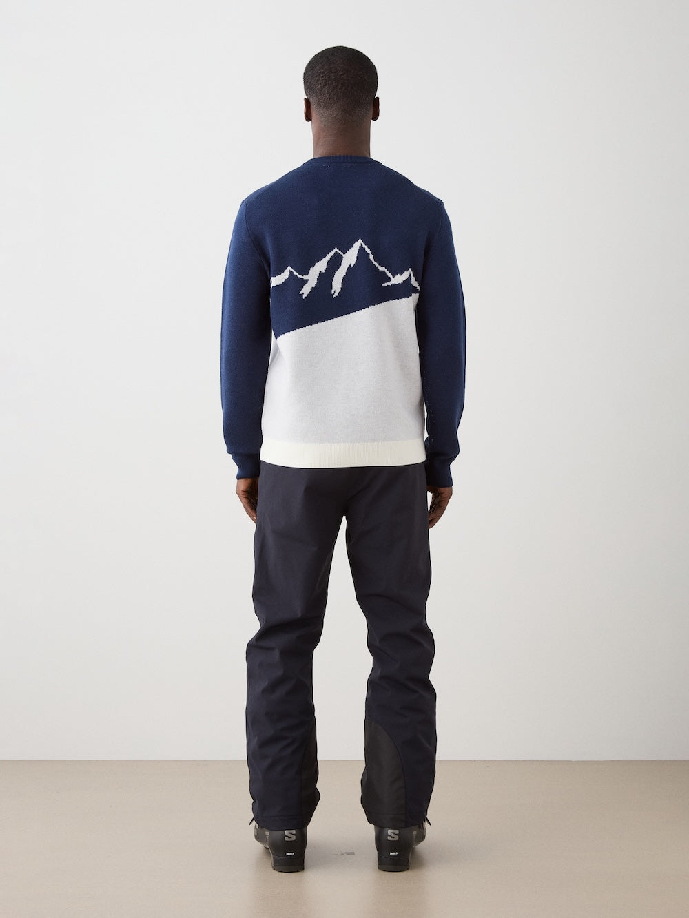 We Norwegians | FW25 | Mens Afterski Crewneck_Navy blue | Model wearing a knitted sweater with an après ski design at the front with black ski pants | Shot against an off-white background | Back view