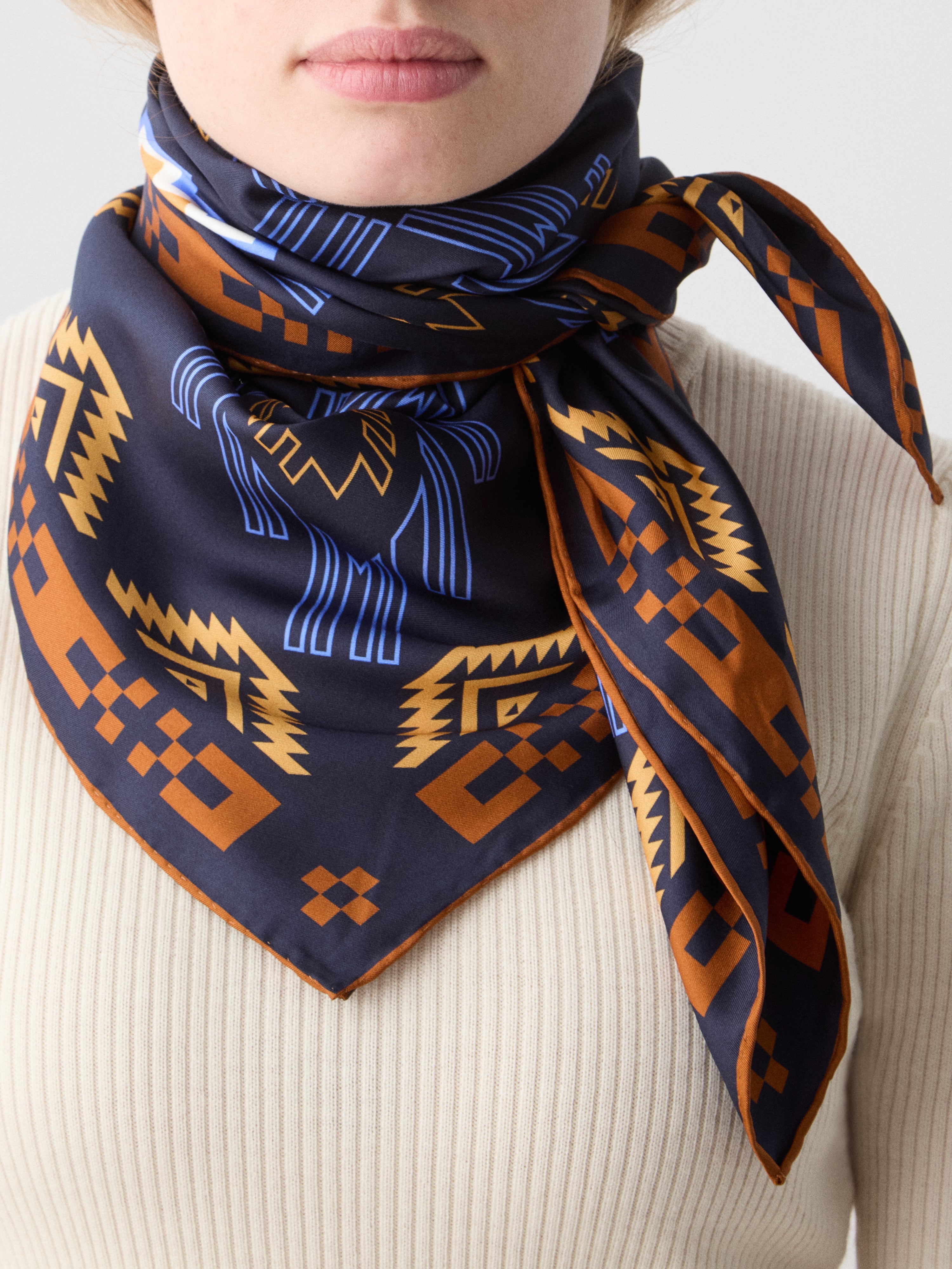 We Norwegians | FW25 | Pioneer Silk Scarf_Navy blue | Female model wearing the scarf with printed motifs inspired by Indigenous American art over a light top | Shot against an off-white background