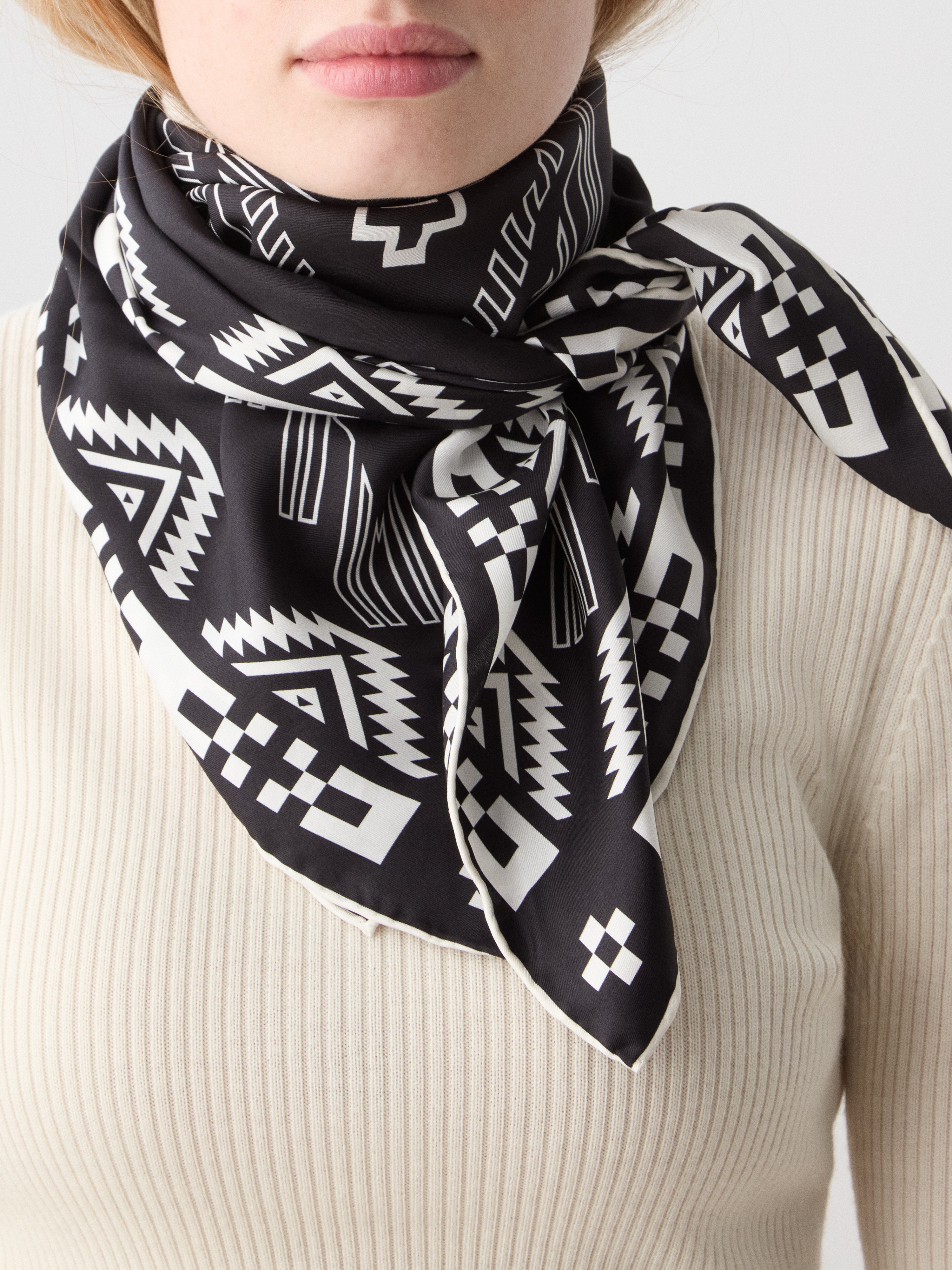 We Norwegians | FW25 | Pioneer Silk Scarf_Black | Female model wearing the scarf with printed motifs inspired by Indigenous American art over a light top | Shot against an off-white background