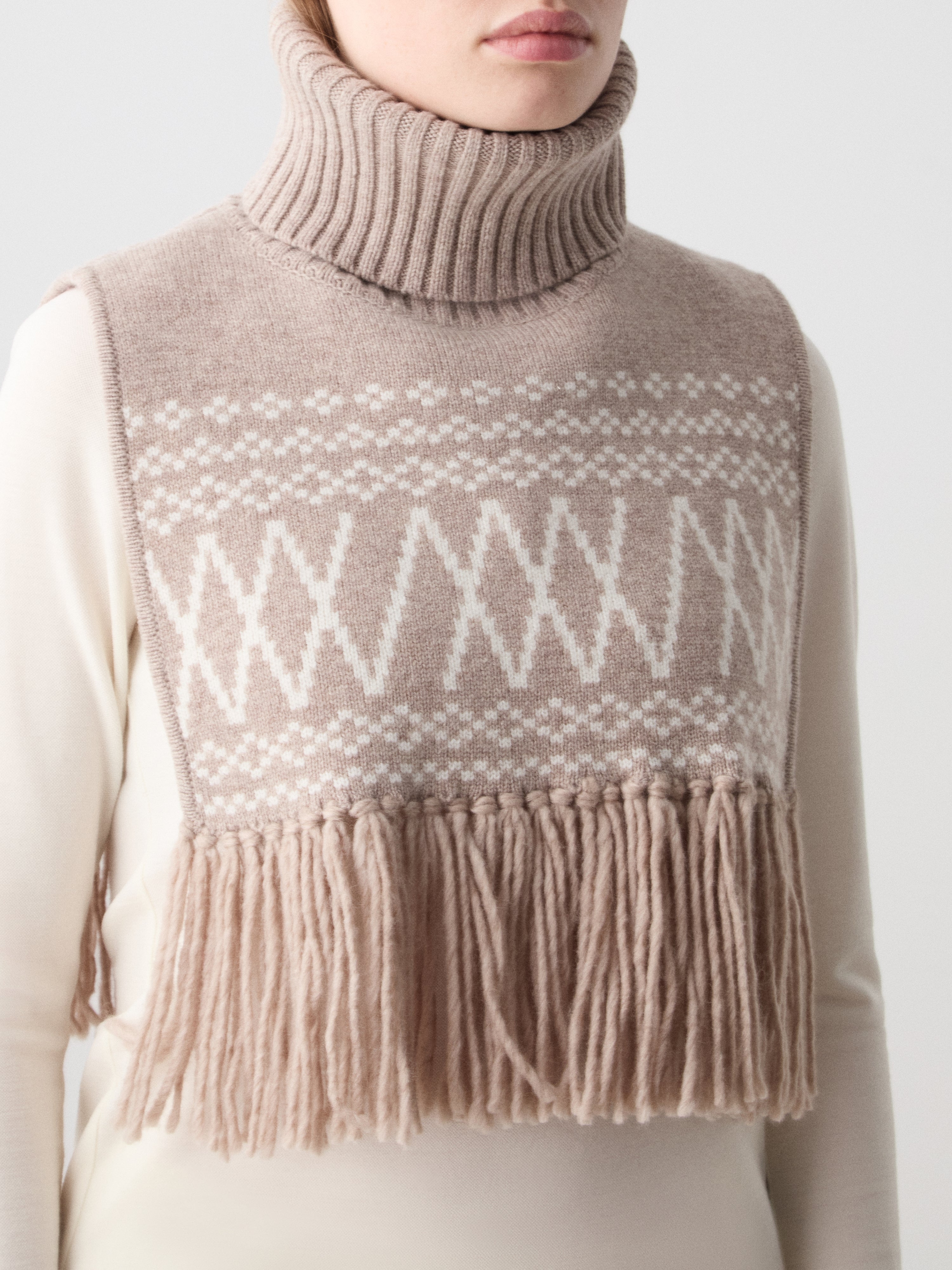We Norwegians | FW25 | Setesdal Bib_Sand | Model is wearing the knitted bib with a turtle neck and fine fringes at the hem over a white sweater, against an off-white background