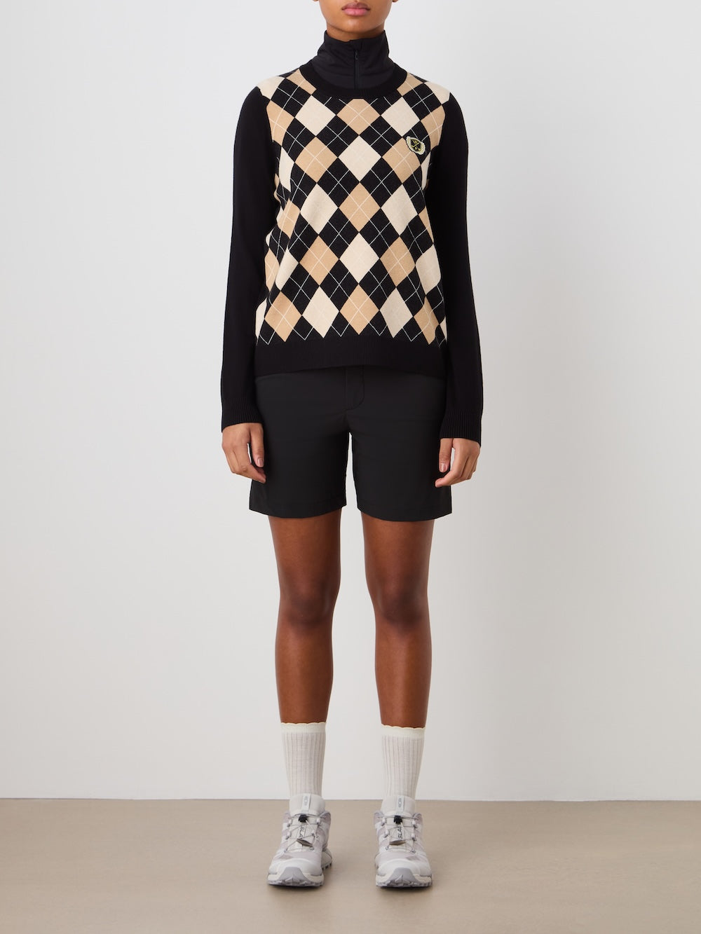 Person wearing the argyle patterned Fairway sweaterblack over a black turtleneck, with black shorts, white socks, and sneakers on a plain background_Front