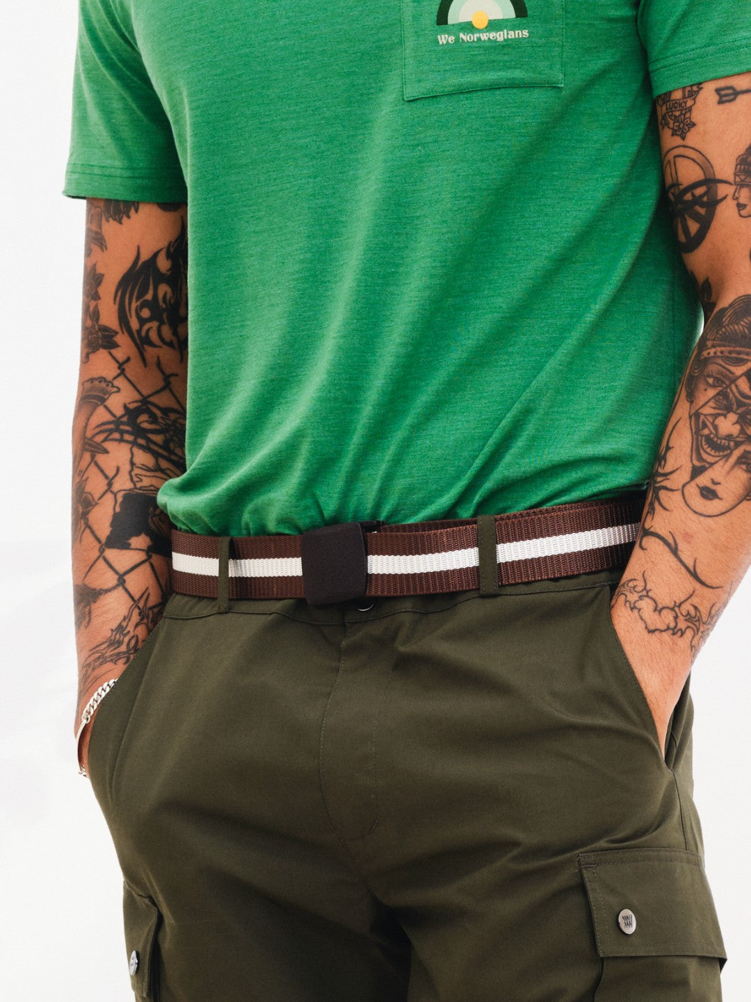 A person is shown from the waist up wearing a green shirt with "We Norwegians" written on the pocket and olive green cargo pants. The individual, adorned with multiple tattoos on both arms, has their hands in their pockets, stylishly showcasing the adjustable Vidda Belt by We Norwegians.